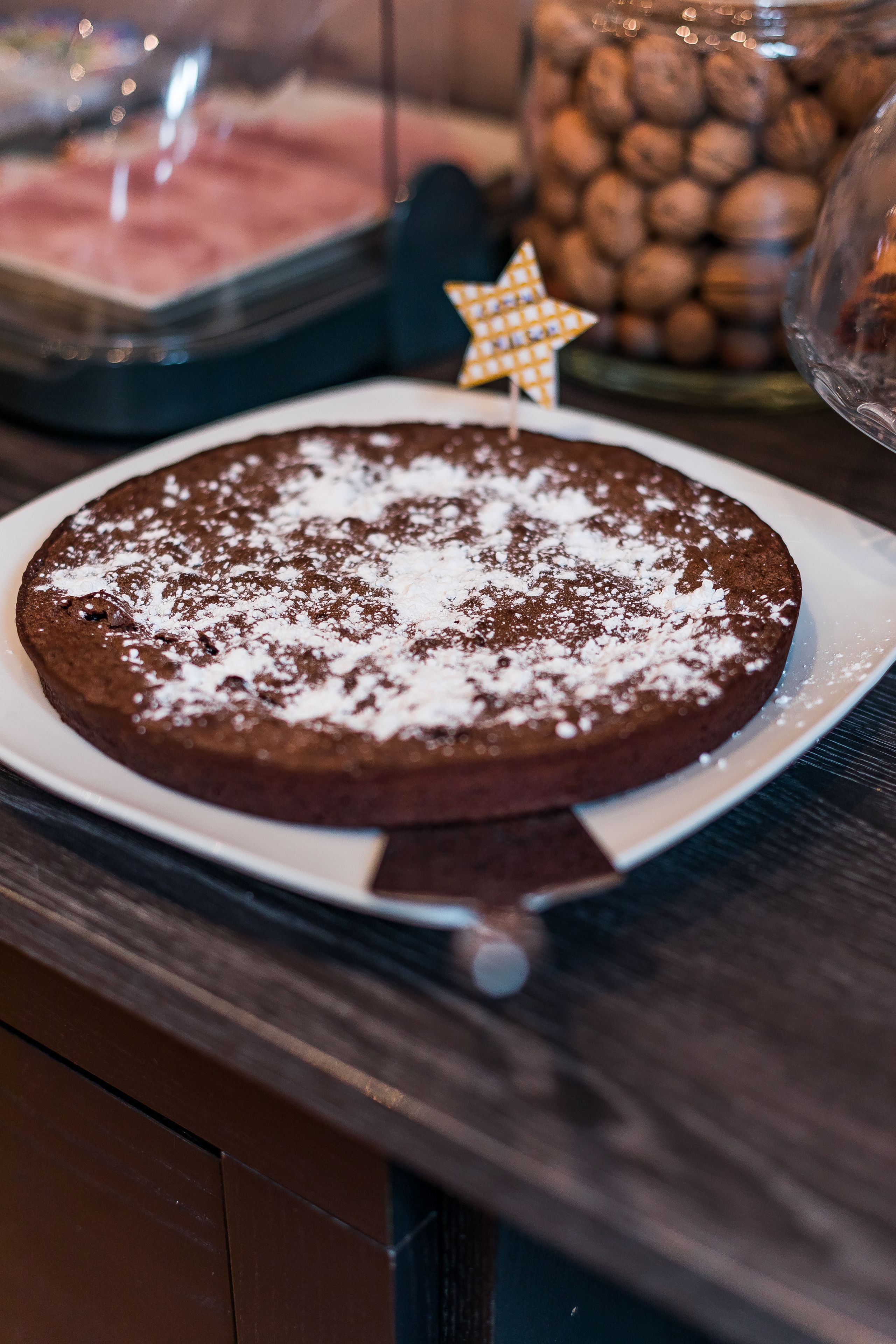 A chocolate cake on a white plate with powdered sugar on top, placed on a wooden table.