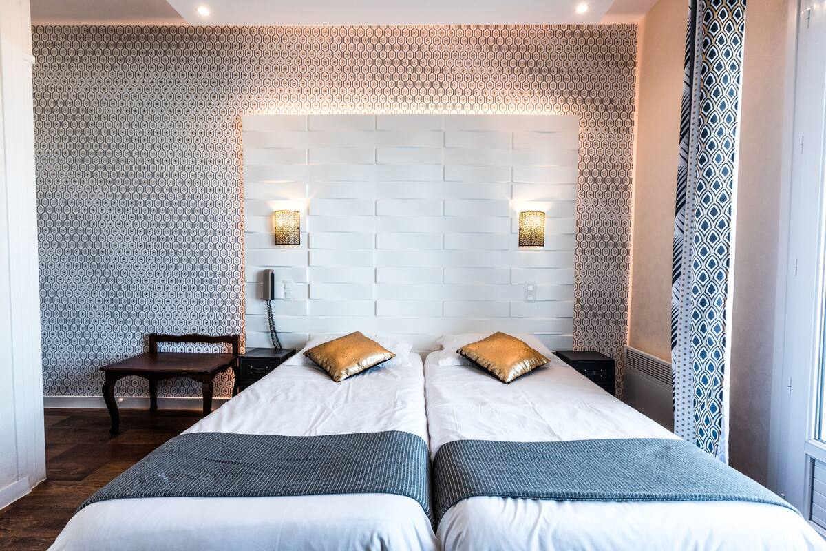 Quarto de hotel com duas camas, um banco de madeira, papel de parede estampado e cabeceira com padrão de tijolos.