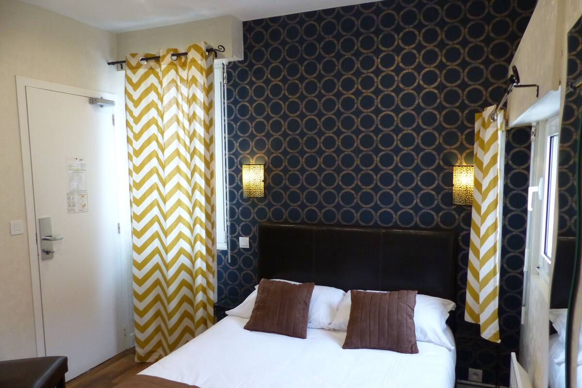 Quarto de hotel com uma cama, um banco, papel de parede estampado e duas luminárias fixadas na parede.