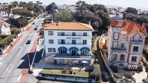Un bâtiment à deux étages, sur lequel on peut lire l’inscription « HÔTEL BELLE-VUE ». Il est doté d’un toit recouvert de tuiles rouges, d’une entrée centrale surmontée d’un auvent, et d’un petit espace extérieur aménagé avec des tables et des chaises.