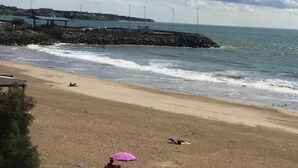 A beach with a rocky breakwater, a person standing near a pole, and another person sitting under a pink umbrella. - Appart Hotel Beau Rivage (Royan)