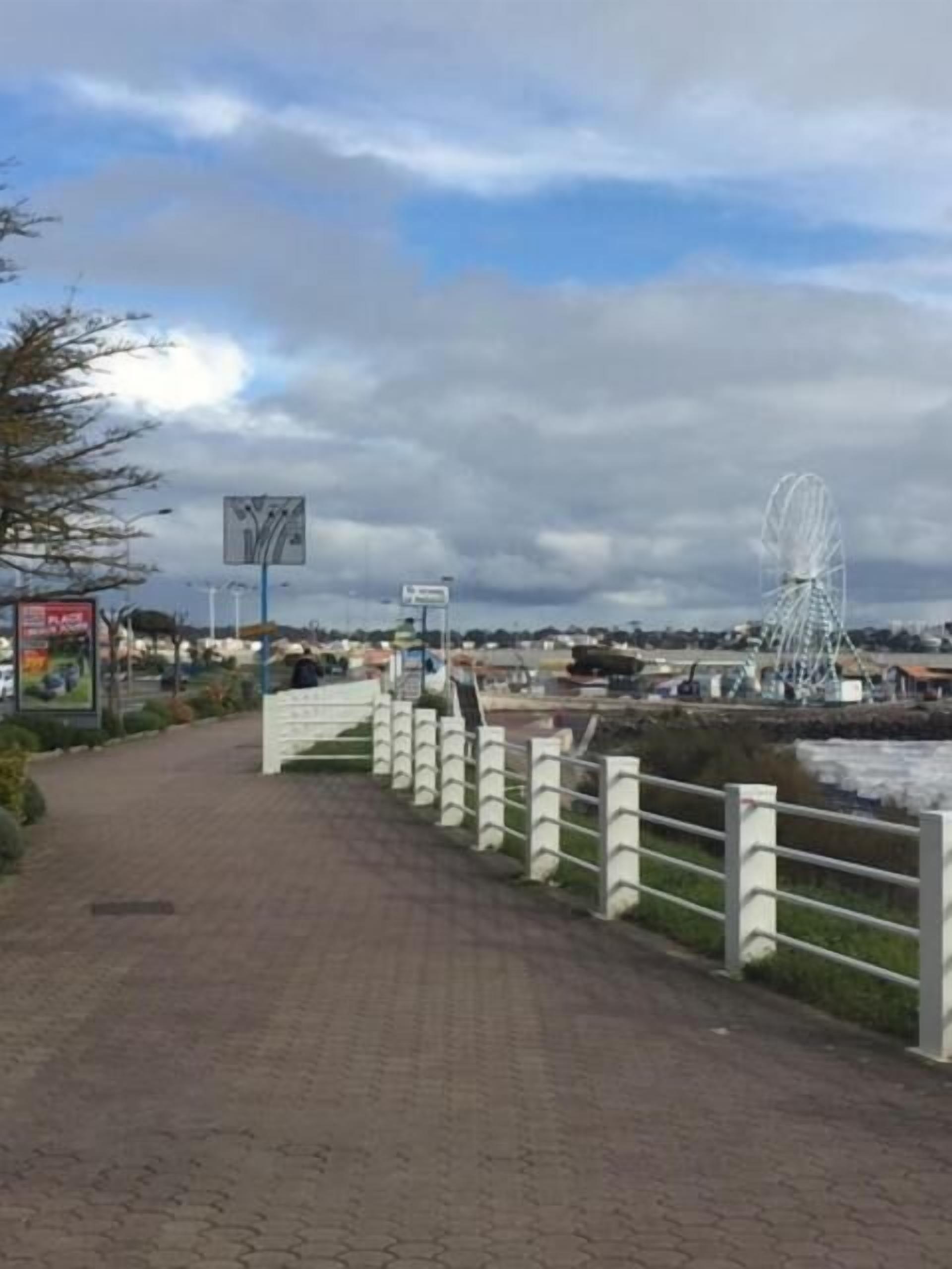 A paved walkway with a white fence, a Ferris wheel, and a cloudy sky.