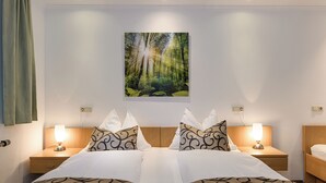 Une chambre d’hôtel avec un lit, deux tables de chevet avec des lampes, un tableau représentant une forêt accroché au mur, et une fenêtre avec des rideaux.