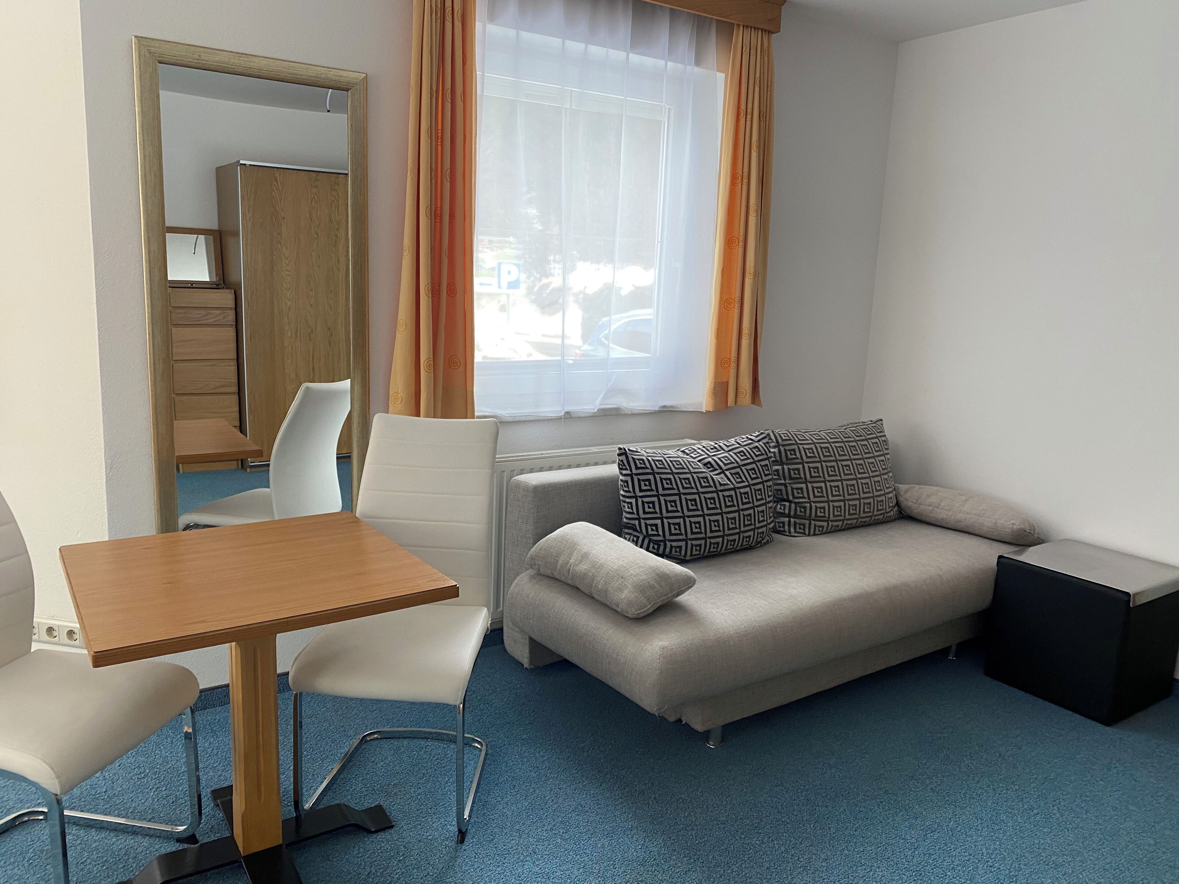 A room with a wooden table, two white chairs, a grey sofa with patterned pillows, a small black side table, and a window with orange curtains.