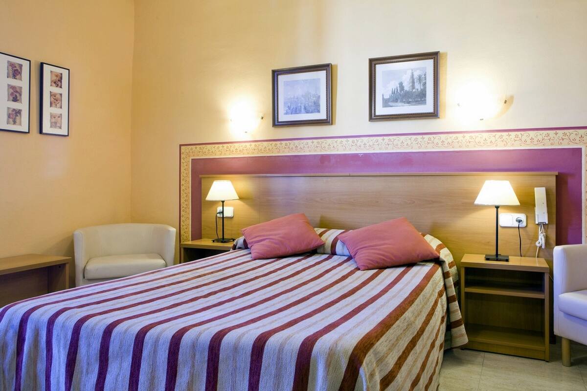 A hotel room with a bed, two bedside tables with lamps, two chairs, and two framed pictures on the wall.
