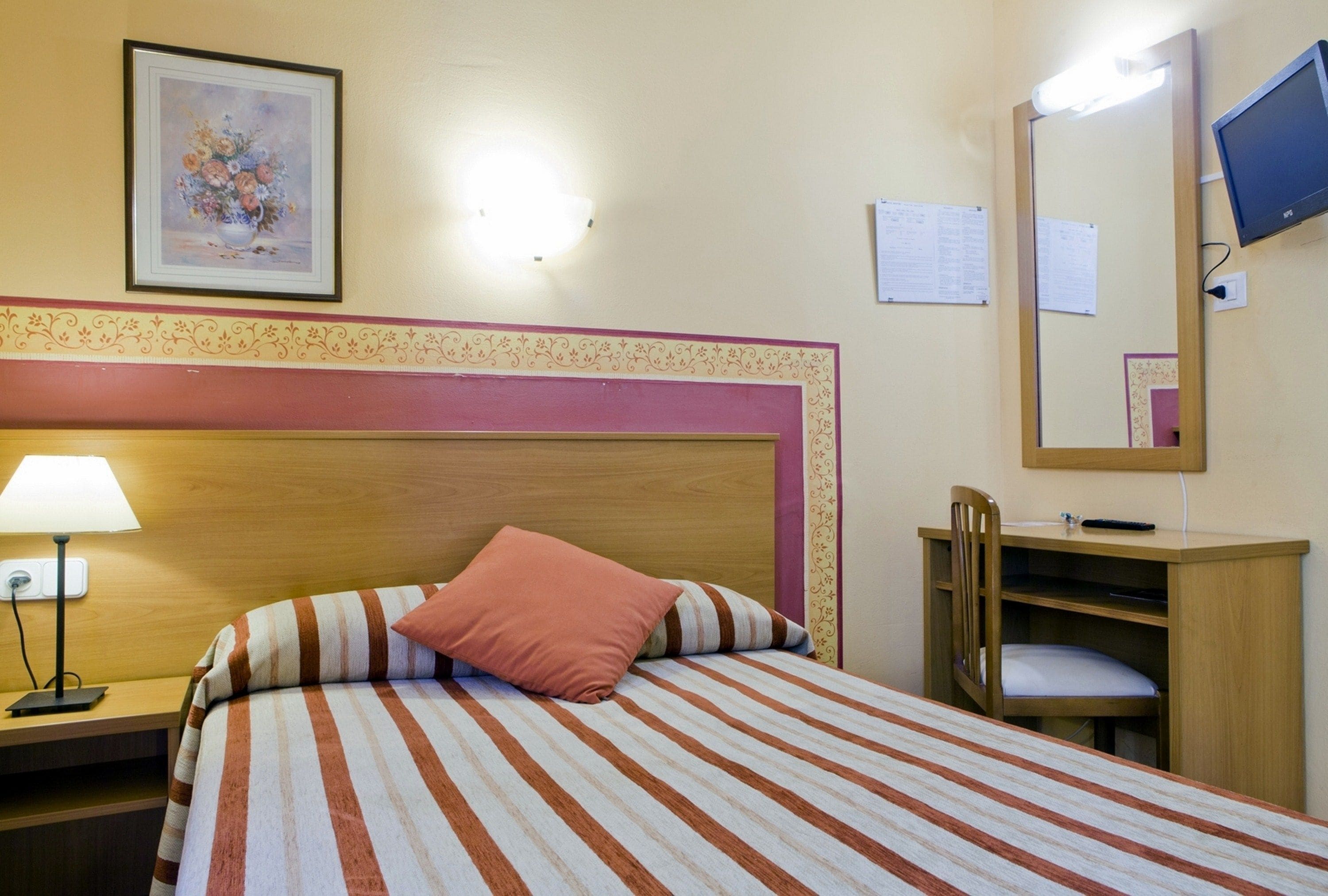 Une chambre d’hôtel comprenant un lit, une table de chevet, une lampe, une tête de lit en bois, un miroir, un bureau et une chaise.
