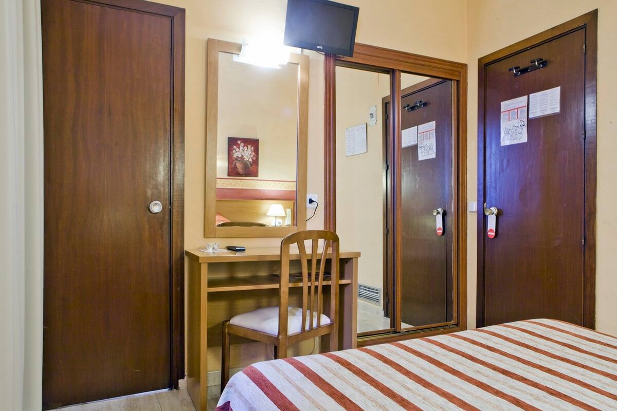 A hotel room with a bed, a desk with a chair, a mirror, and two doors.