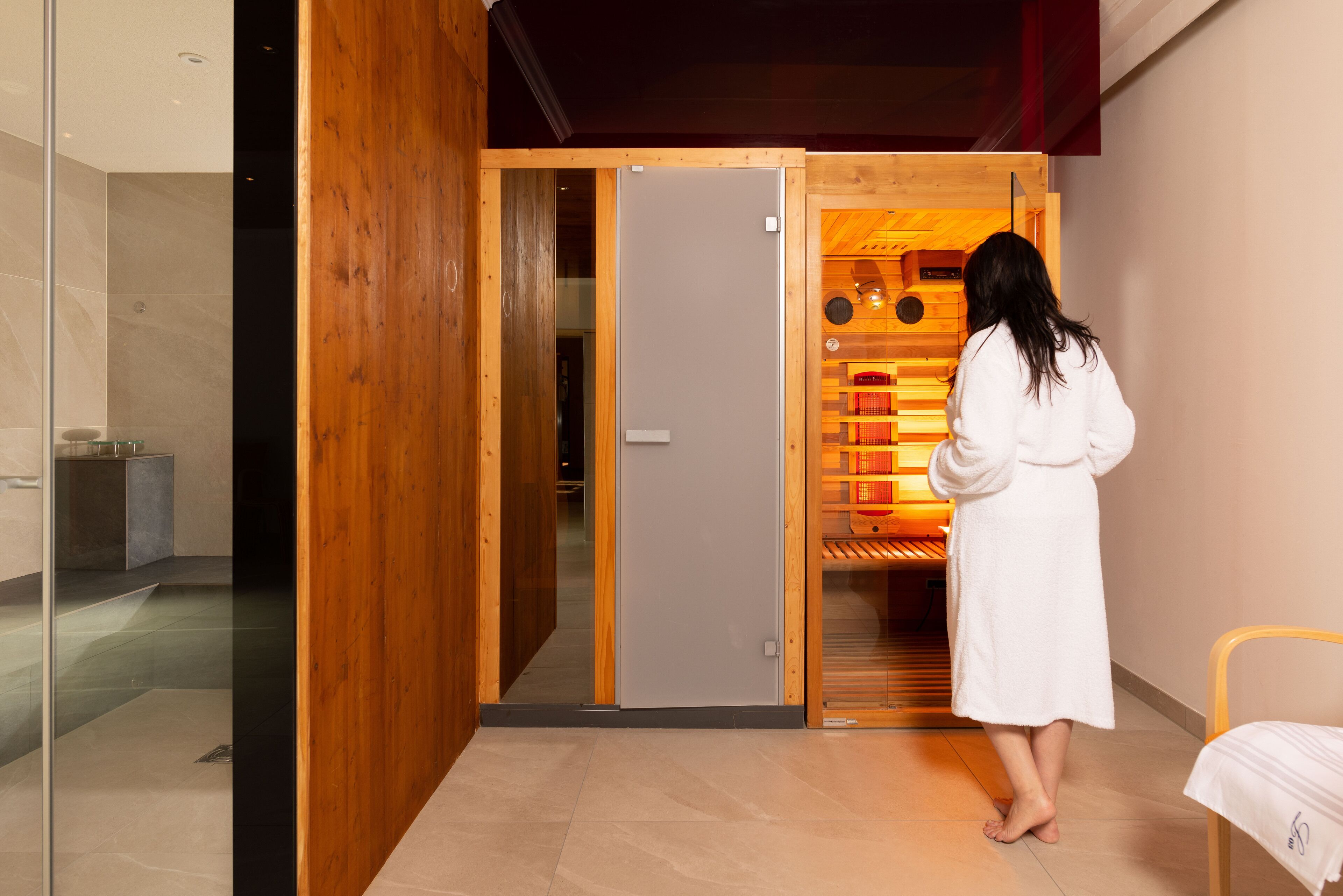 A person in a white robe standing in a room with a wooden door leading to a sauna area.