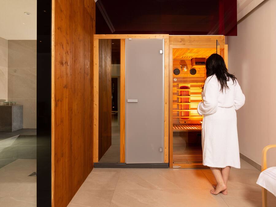 A person in a white robe standing in a room with a wooden door leading to a sauna area.