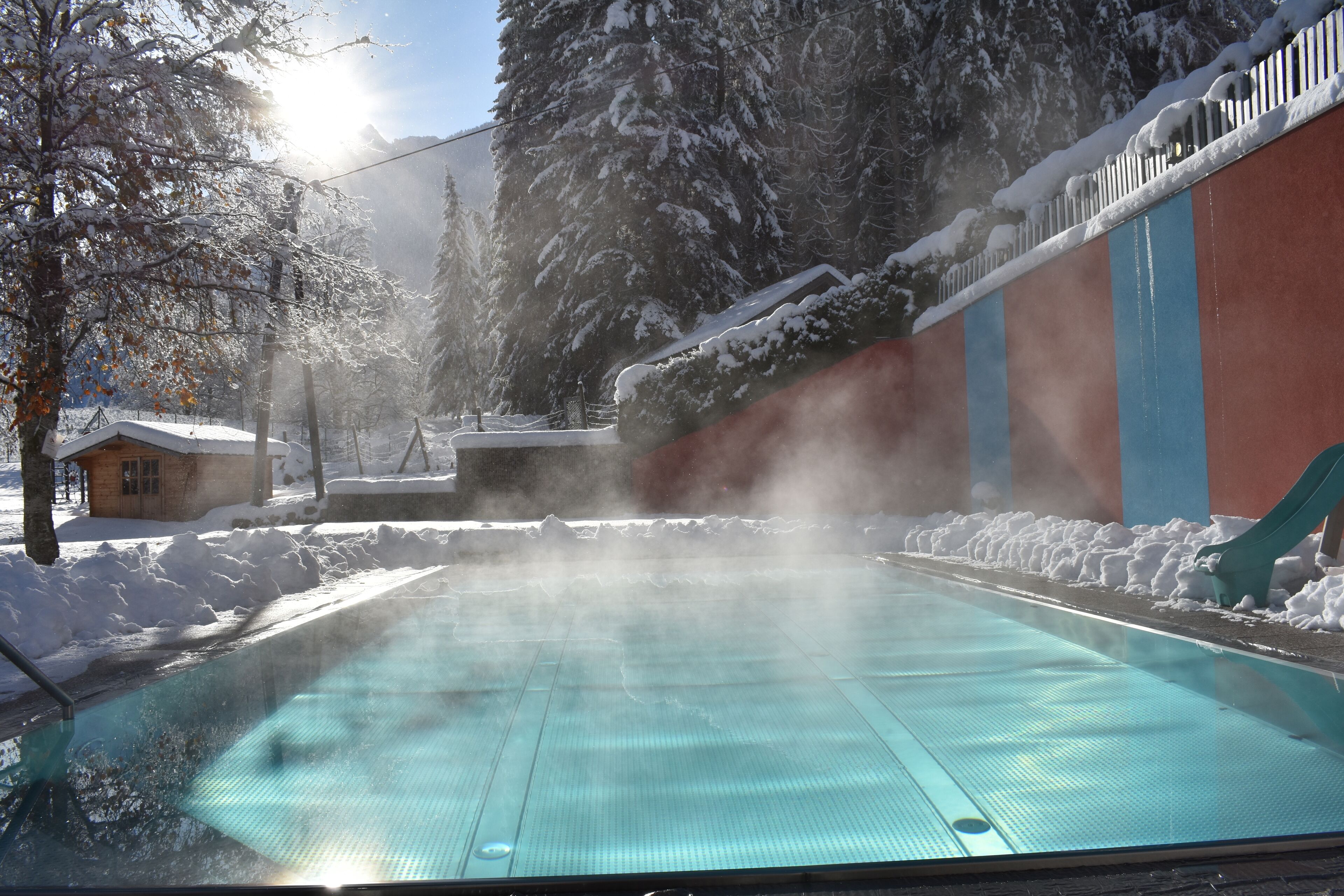 Une piscine d’un bleu éclatant, entourée d’arbres couverts de neige, et un bâtiment à la façade rouge et bleue.