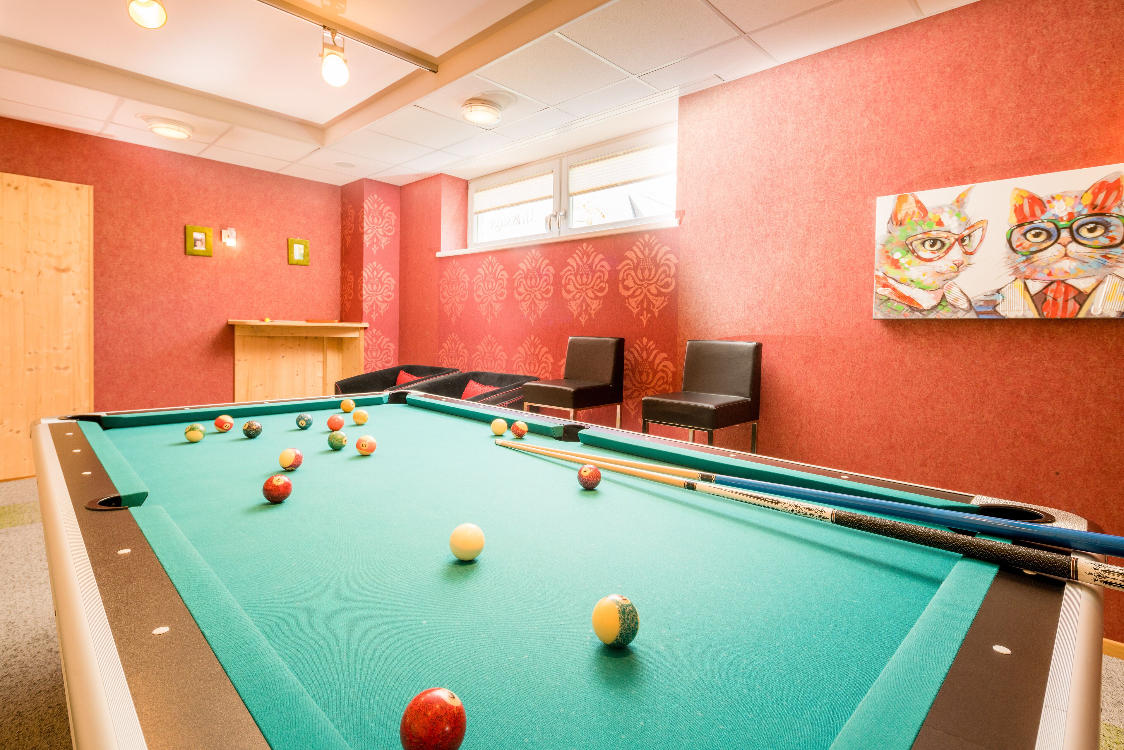 A pool table in a room with red walls, a window, and a painting of cats.