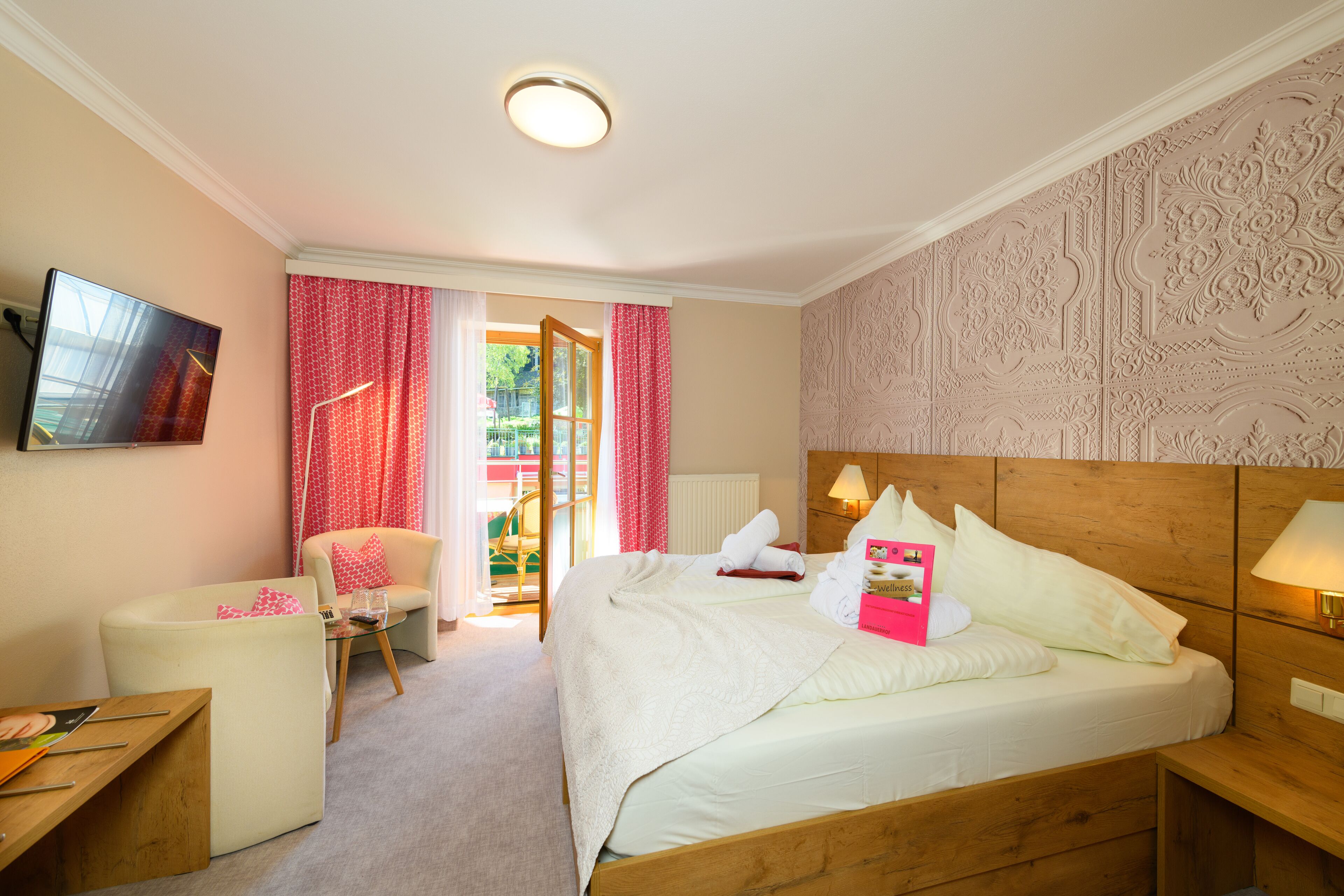 A hotel room with a wooden headboard, a bed with white linens, a small table with a lamp, a chair, and a television on the wall.
