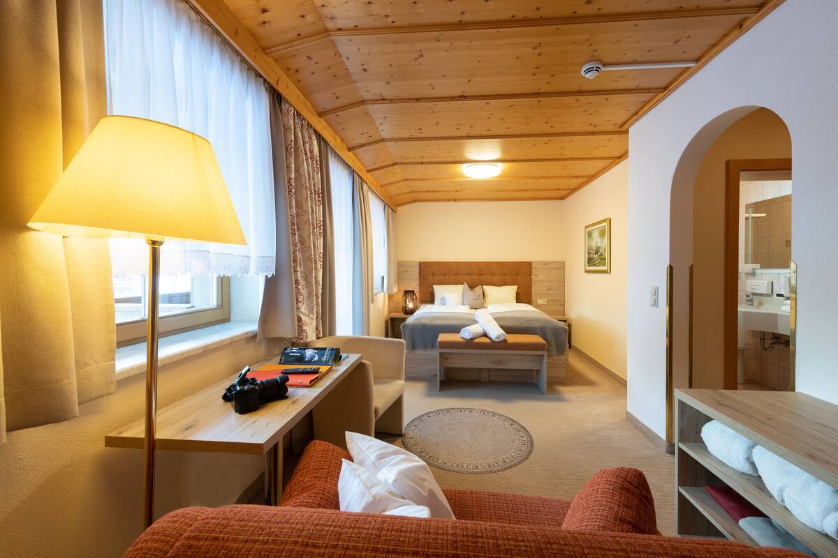 Une chambre d’hôtel avec un lit, un bureau doté d’une lampe, une chaise, un canapé et une salle de bain visible à travers une arche.