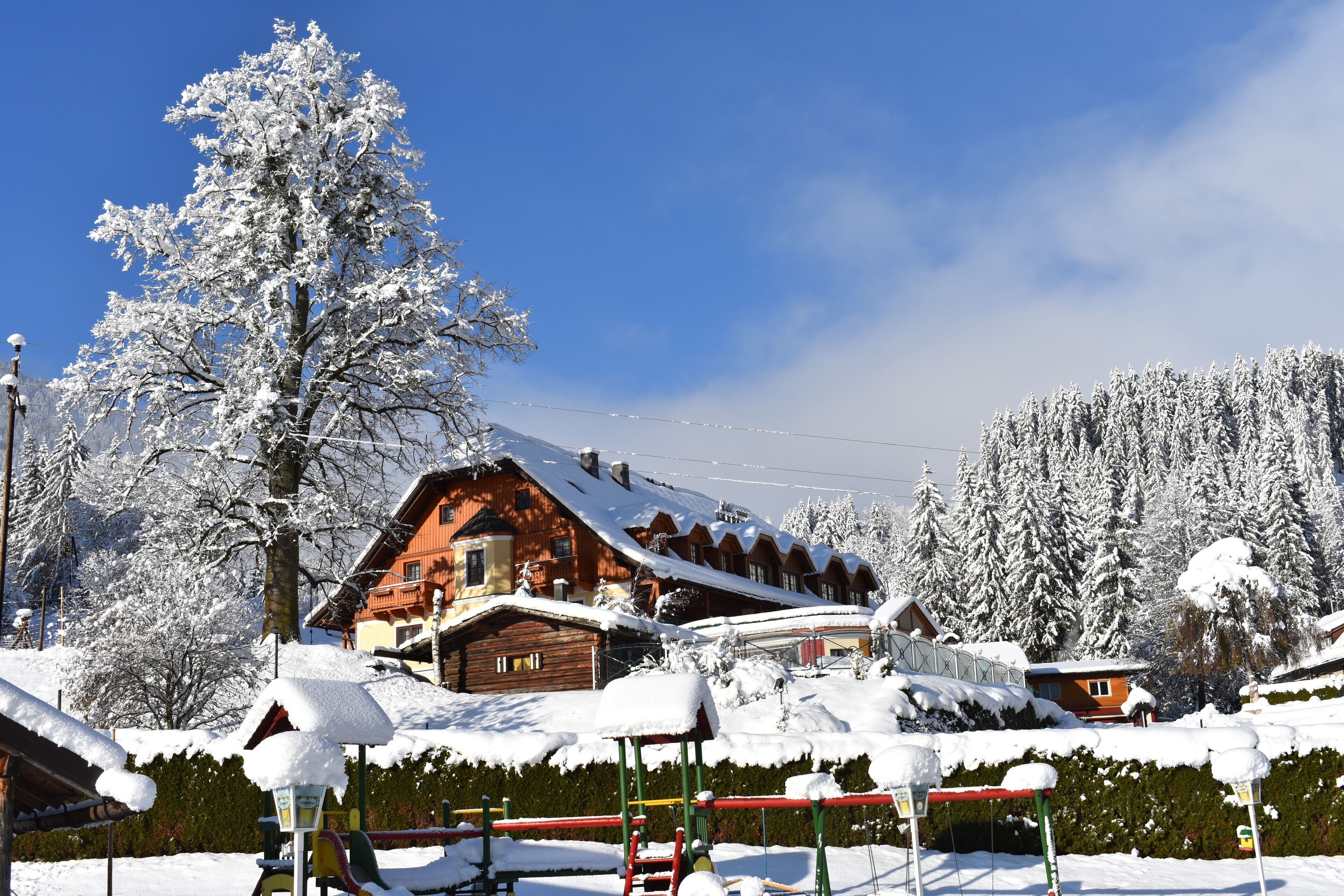 A snow-covered wooden chalet with a sloped roof, surrounded by snow-laden trees and a clear blue sky.