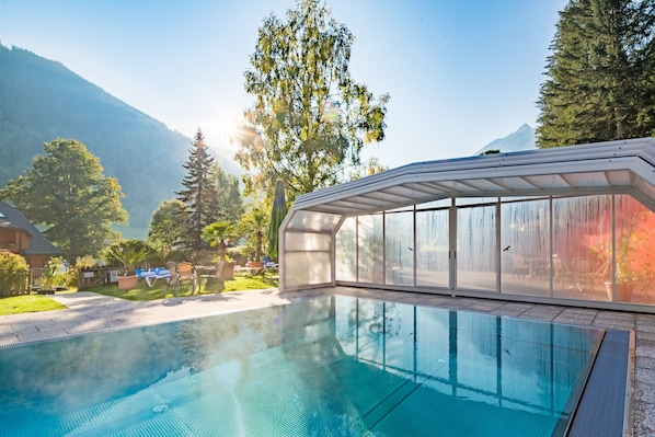 Una piscina con cerramiento de vidrio, rodeada de árboles y una montaña al fondo.