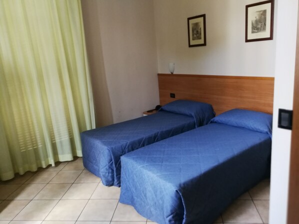 Une chambre avec deux lits simples, une tête de lit en bois, un rideau et deux tableaux encadrés au mur.