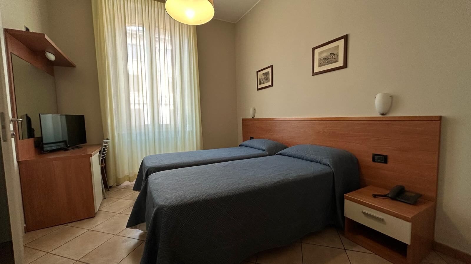 Habitación de hotel con dos camas, un escritorio, televisor y ventana con cortinas.