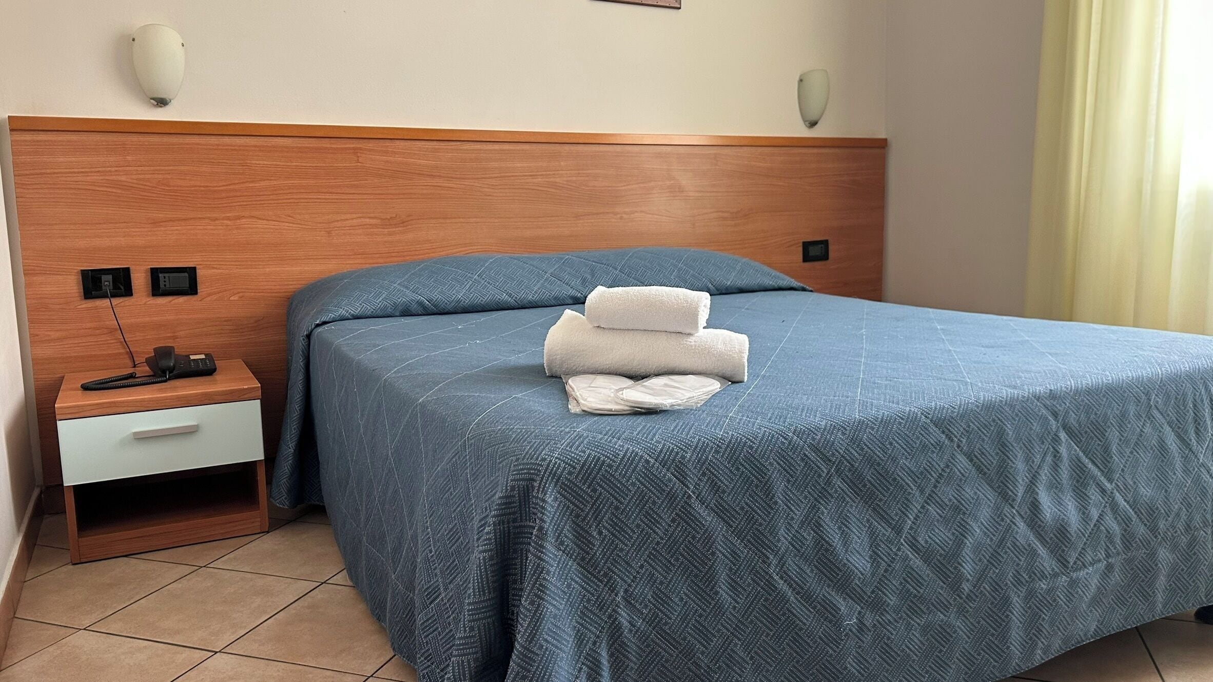A hotel room with a wooden headboard, a blue bedspread, a bedside table with a phone, and a wall-mounted light fixture.