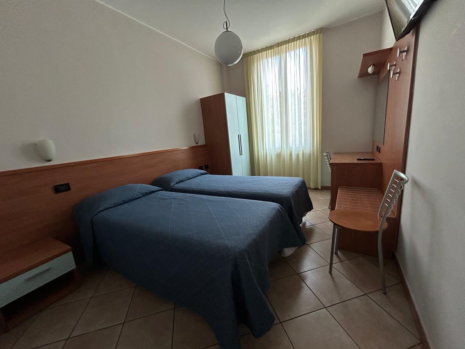 A hotel room with two beds, a desk, a chair, a wardrobe, and a window with curtains.