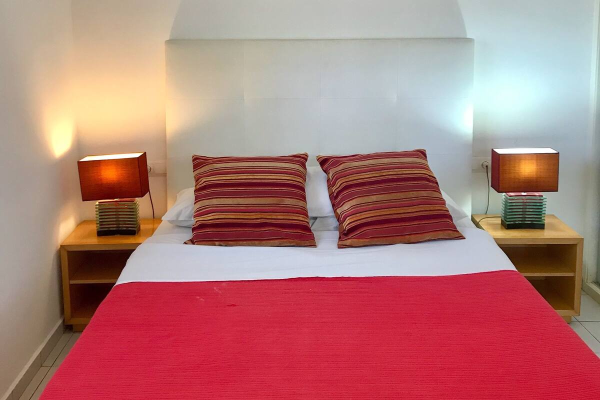 Una cama bien hecha con una colcha roja y almohadas a rayas, flanqueada por dos mesitas de noche con lámparas, y un cuadro abstracto y colorido sobre la cama.