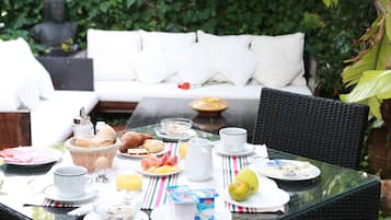 Mesa de desayuno al aire libre con diversos alimentos, un sofá blanco al fondo y follaje verde.