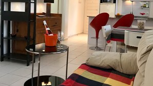 A modern living room with a sofa, a dining table, and red chairs. There is a kitchen in the background with white cabinets and a microwave. - Hotel los Globos (Sitges)