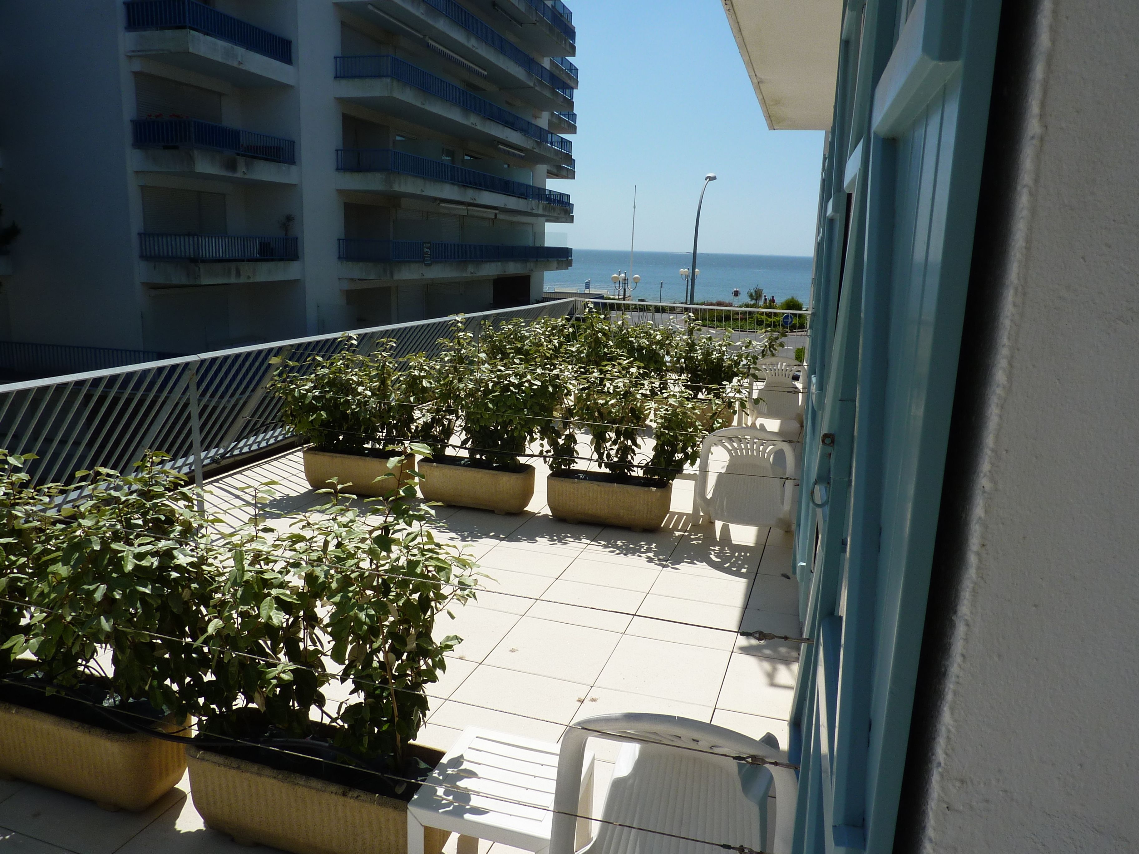 A balcony with potted plants and white chairs, overlooking a building and the sea.