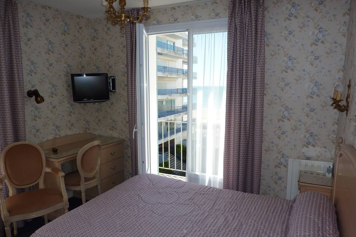Habitación de hotel con cama, escritorio con dos sillas, televisor y balcón con barandillas azules.