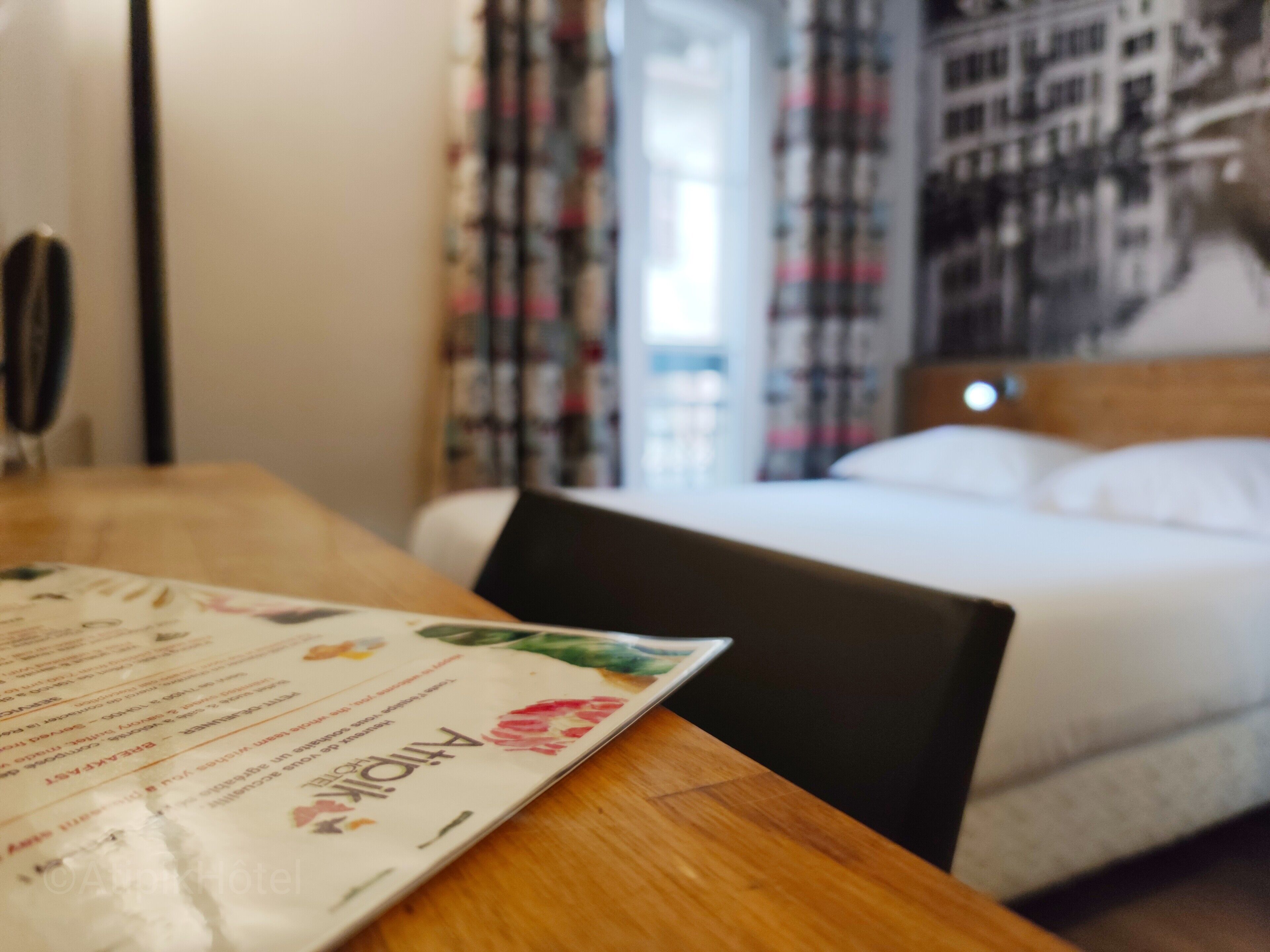 Hotellihuone, jossa on sänky, puinen pöytä, jossa on ruokalista, ja ikkunasta avautuva näkymä kaupunkiin.