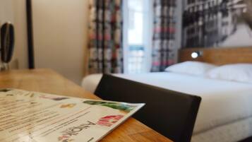 Une chambre d’hôtel avec un lit, une table en bois sur laquelle se trouve un menu, et une vue sur la ville par la fenêtre.