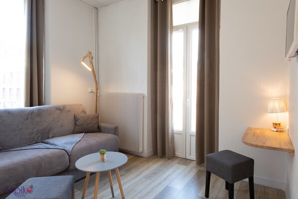 A modern living room with a grey sofa, a small round table with a plant, a wooden bench, a standing lamp, and a radiator. - Atipik Hôtel (Annecy)