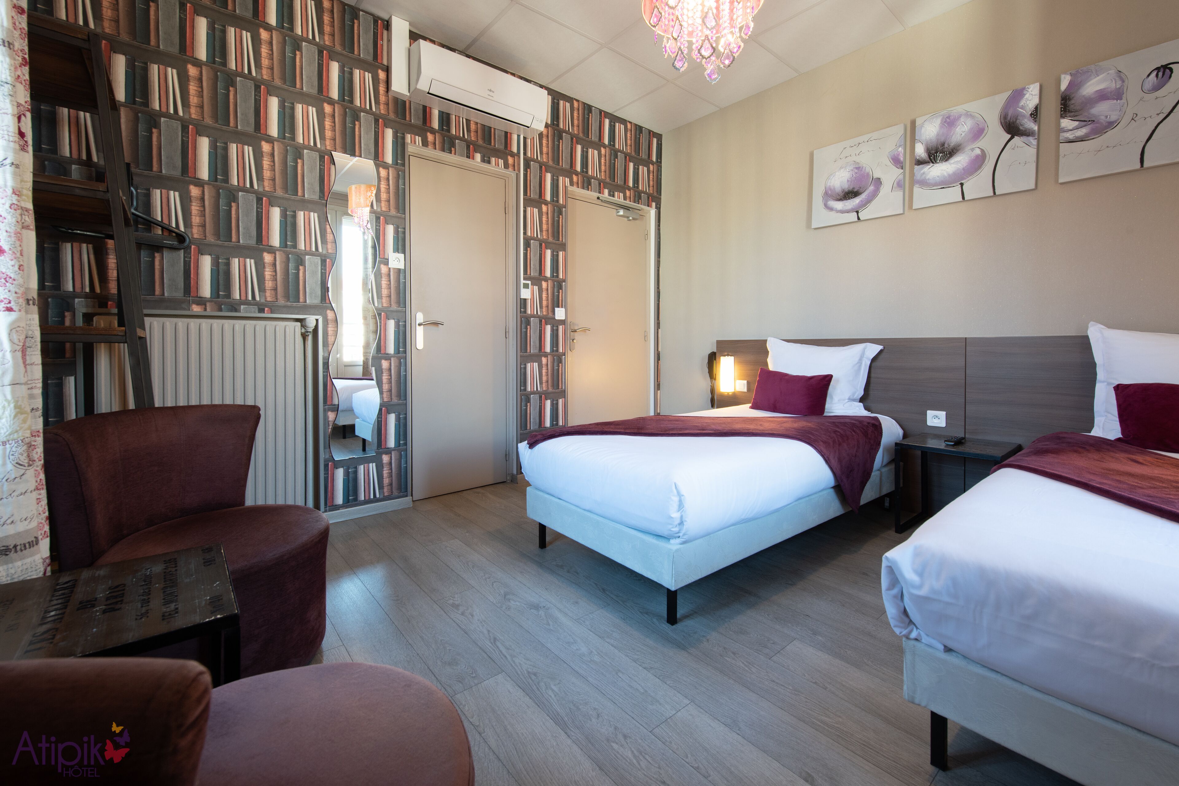 A hotel room with two beds, a bookshelf, a radiator, and a chandelier.