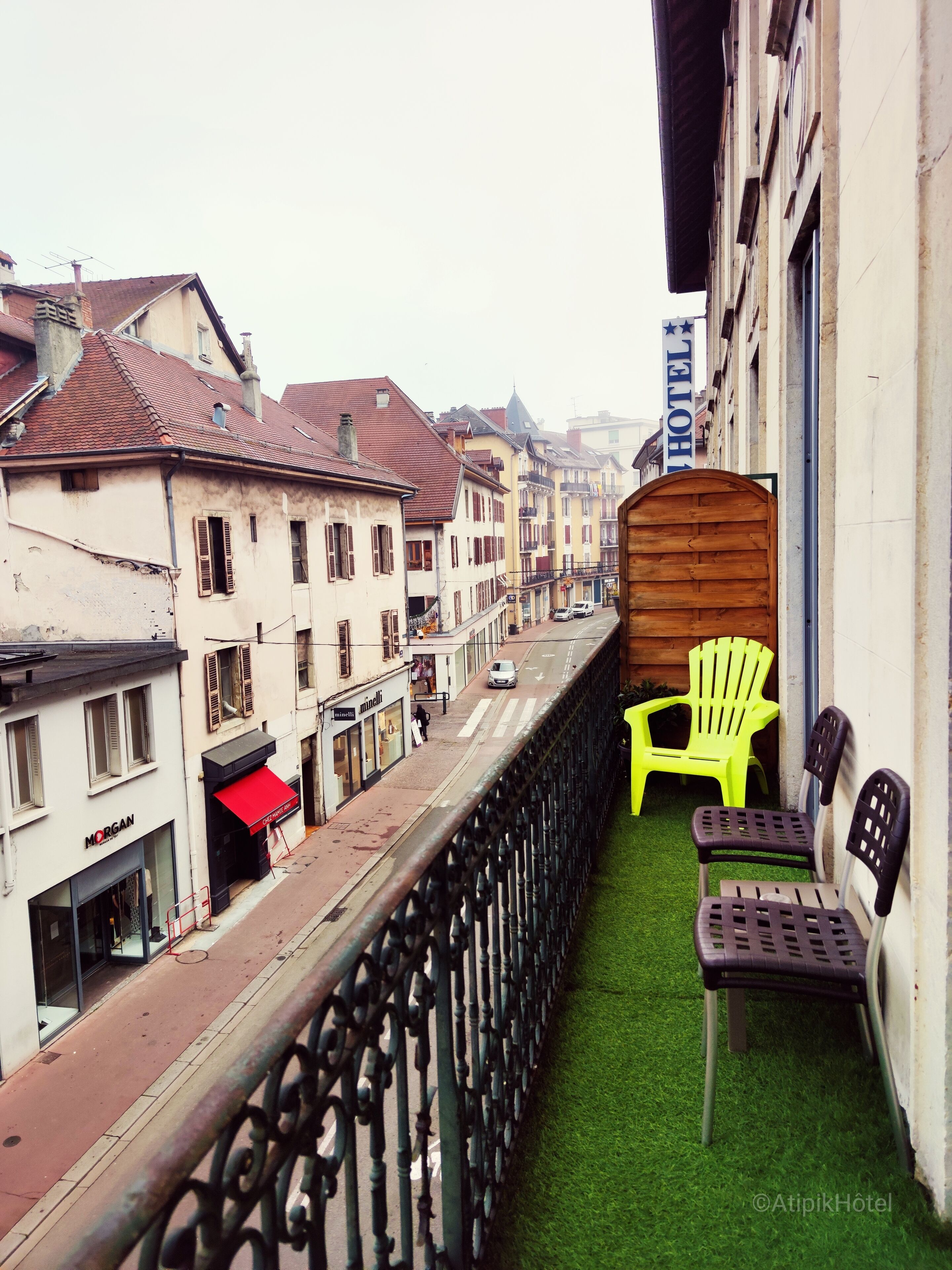 Un balcon avec une barrière en bois, deux chaises en plastique et une vue sur une rue bordée d’immeubles et d’une enseigne d’hôtel.