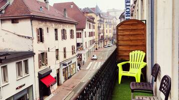Un balcon avec une barrière en bois, deux chaises en plastique et une vue sur une rue bordée d’immeubles et d’une enseigne d’hôtel.