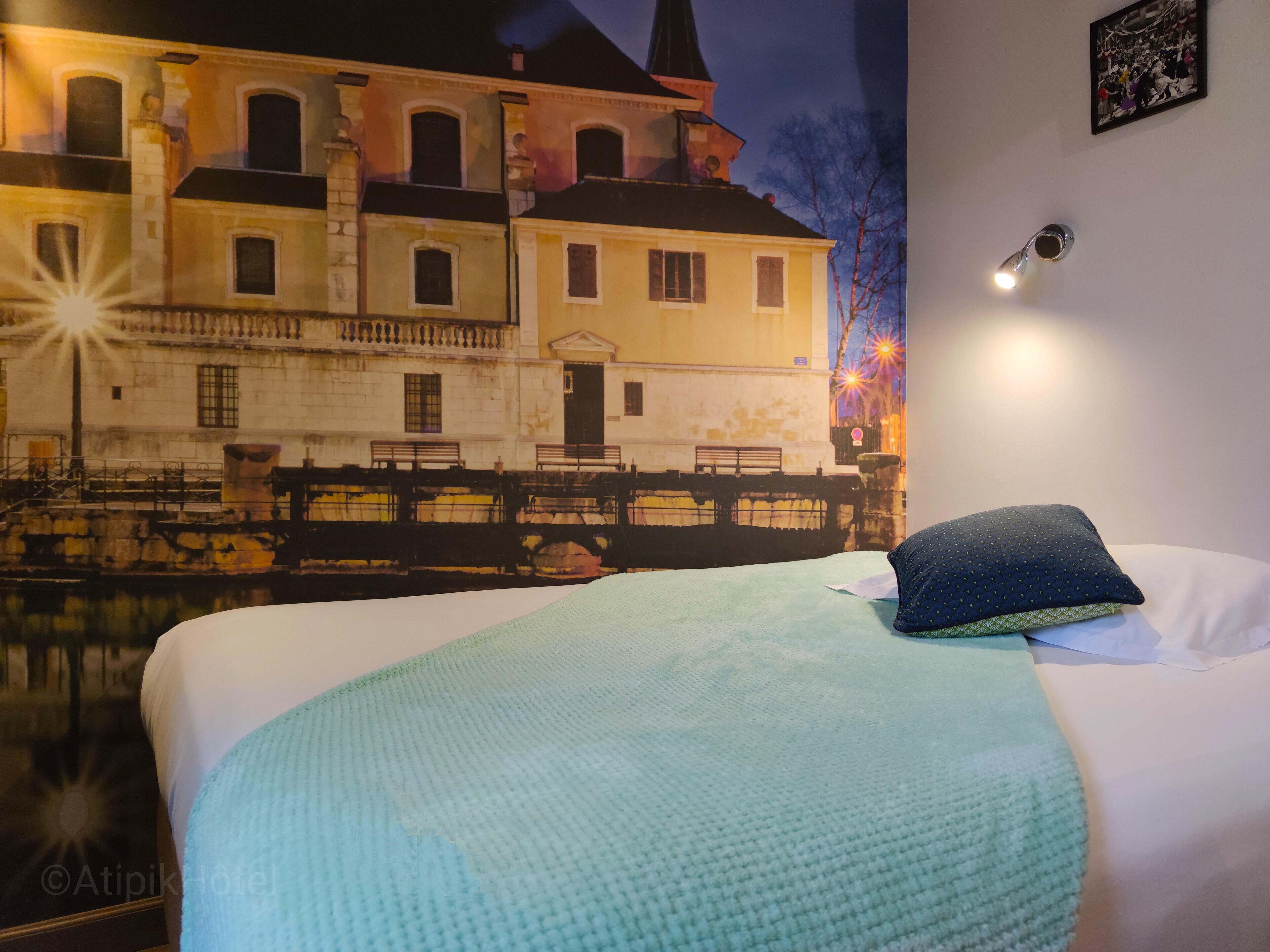 Une chambre d’hôtel avec un lit, une couverture bleue, un oreiller et une fresque murale représentant un bâtiment.