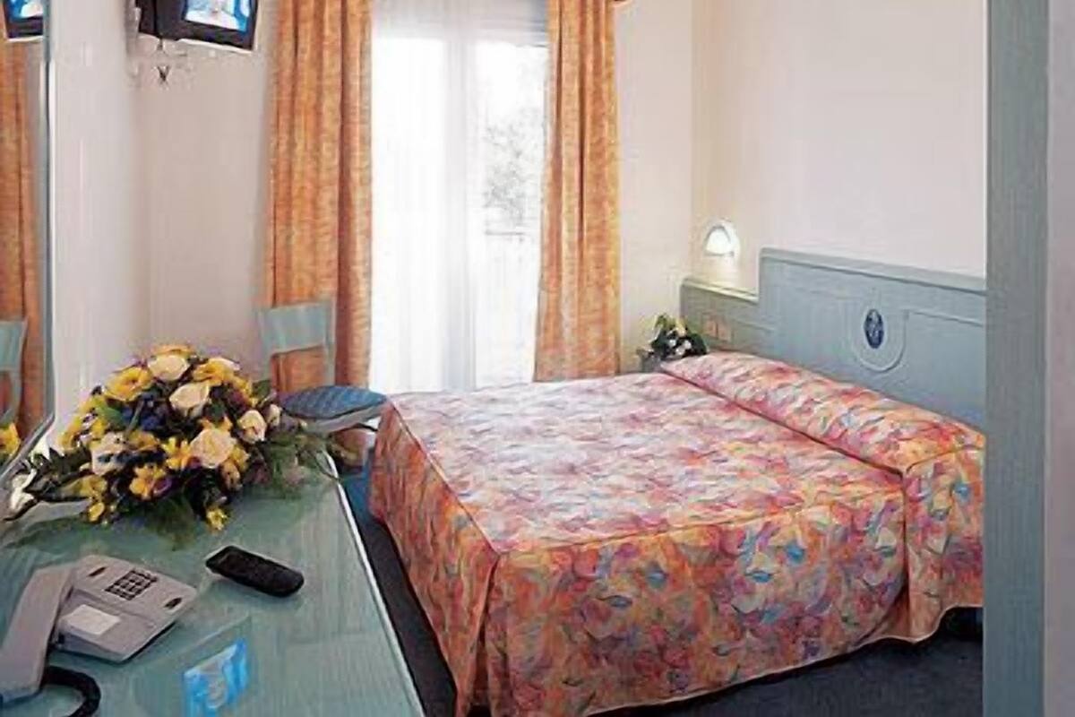 Habitación de hotel con cama, escritorio con teléfono y control remoto, televisor montado en la pared y ventana con cortinas.