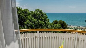 Ein Balkon mit weißen Stühlen, einem kleinen runden Tisch mit Teekanne, Tassen und Gebäck, mit Blick auf einen Strand und ein klares blaues Meer, im Hintergrund grüne Bäume.