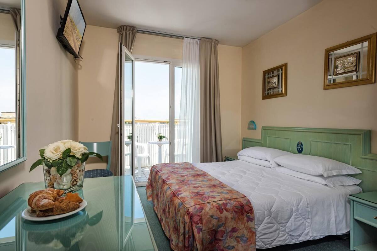 Habitación de hotel con una cama, una mesa con un plato de comida, un jarrón con flores y vistas al balcón exterior.