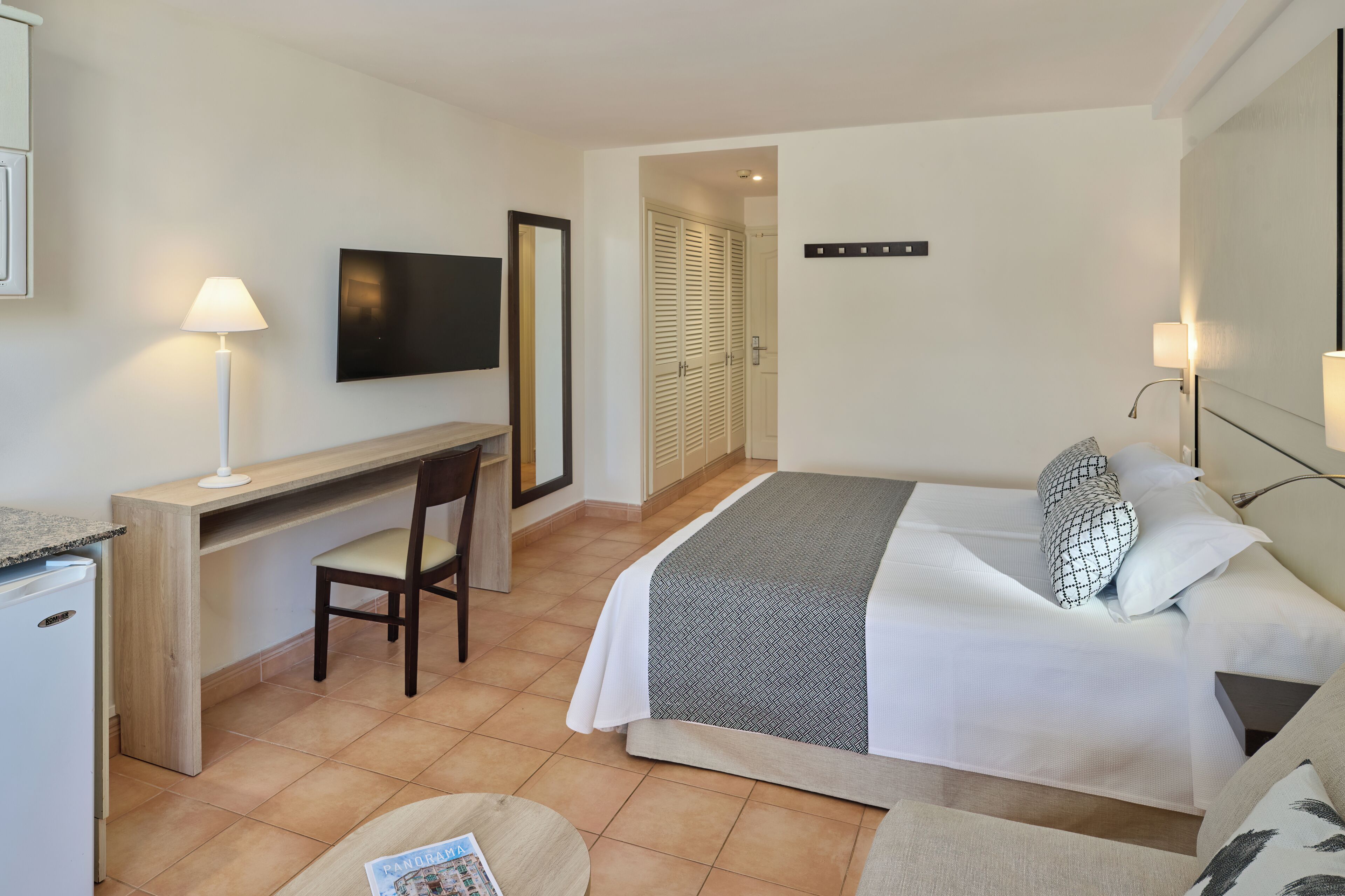 A hotel room with a bed, a desk, a chair, a TV, and a small kitchenette.