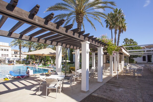An outdoor pool area with lounge chairs and umbrellas, surrounded by palm trees and a pergola structure.