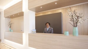 Ein Empfangsschalter mit modernem Design, mit heller Holzoptik und einer dunkel gestalteten Akzentwand. Auf dem Schalter stehen zwei Vasen mit Zweigen.