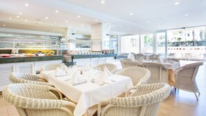 Ein Restaurant mit Korbstühlen und Tischen, gedeckt zum Essen, einem Buffet mit verschiedenen Speisen und großen Fenstern mit Blick ins Freie.