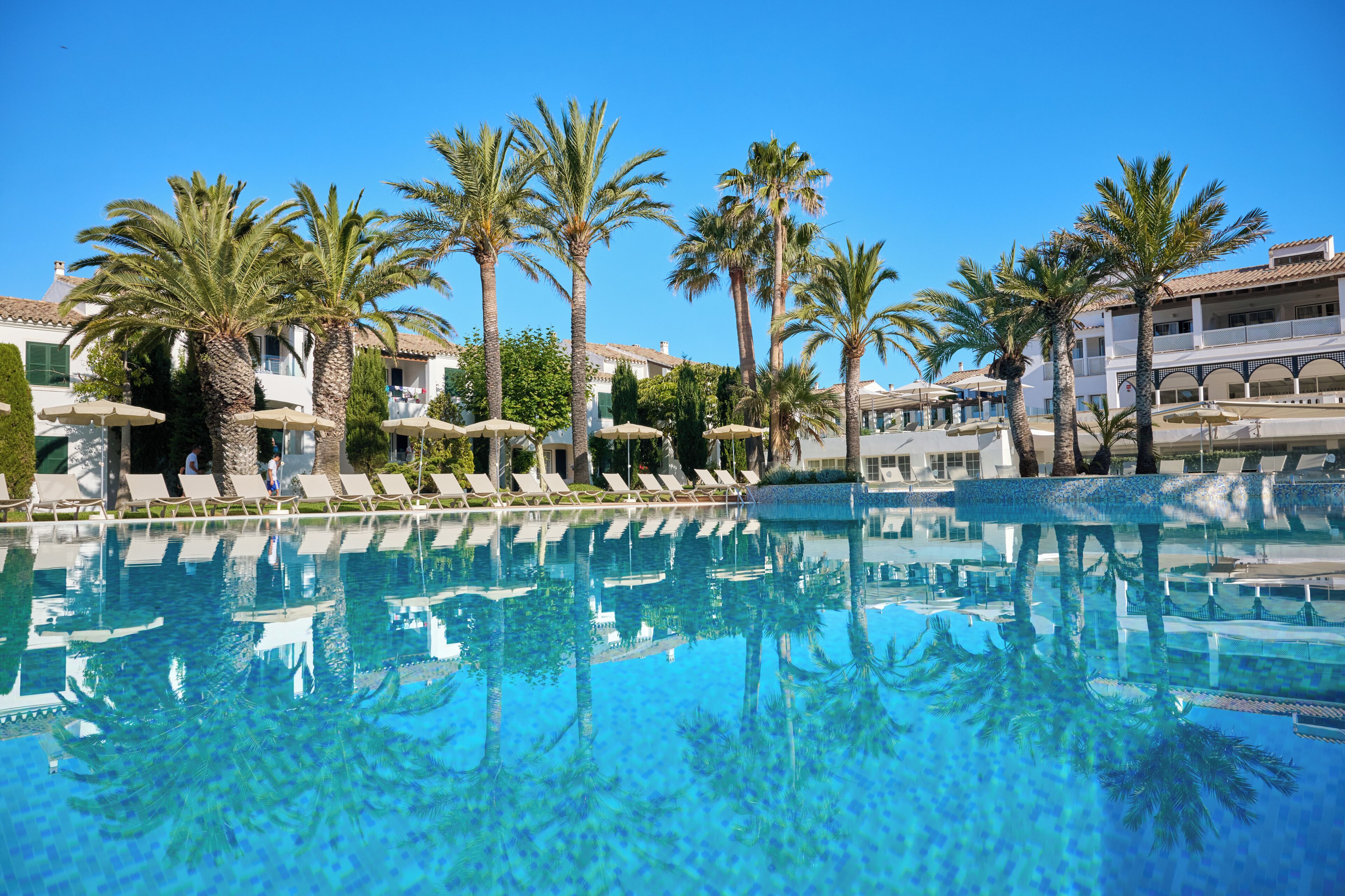 Una piscina azul cristalina con sillas de descanso y sombrillas, rodeada de palmeras y edificios con balcones.
