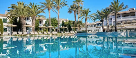 Una piscina azul cristalina con sillas de descanso y sombrillas, rodeada de palmeras y edificios con balcones.
