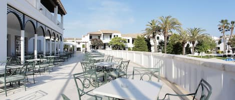 An outdoor cafe area with multiple tables and chairs, a white building with arches, and palm trees in the background.
