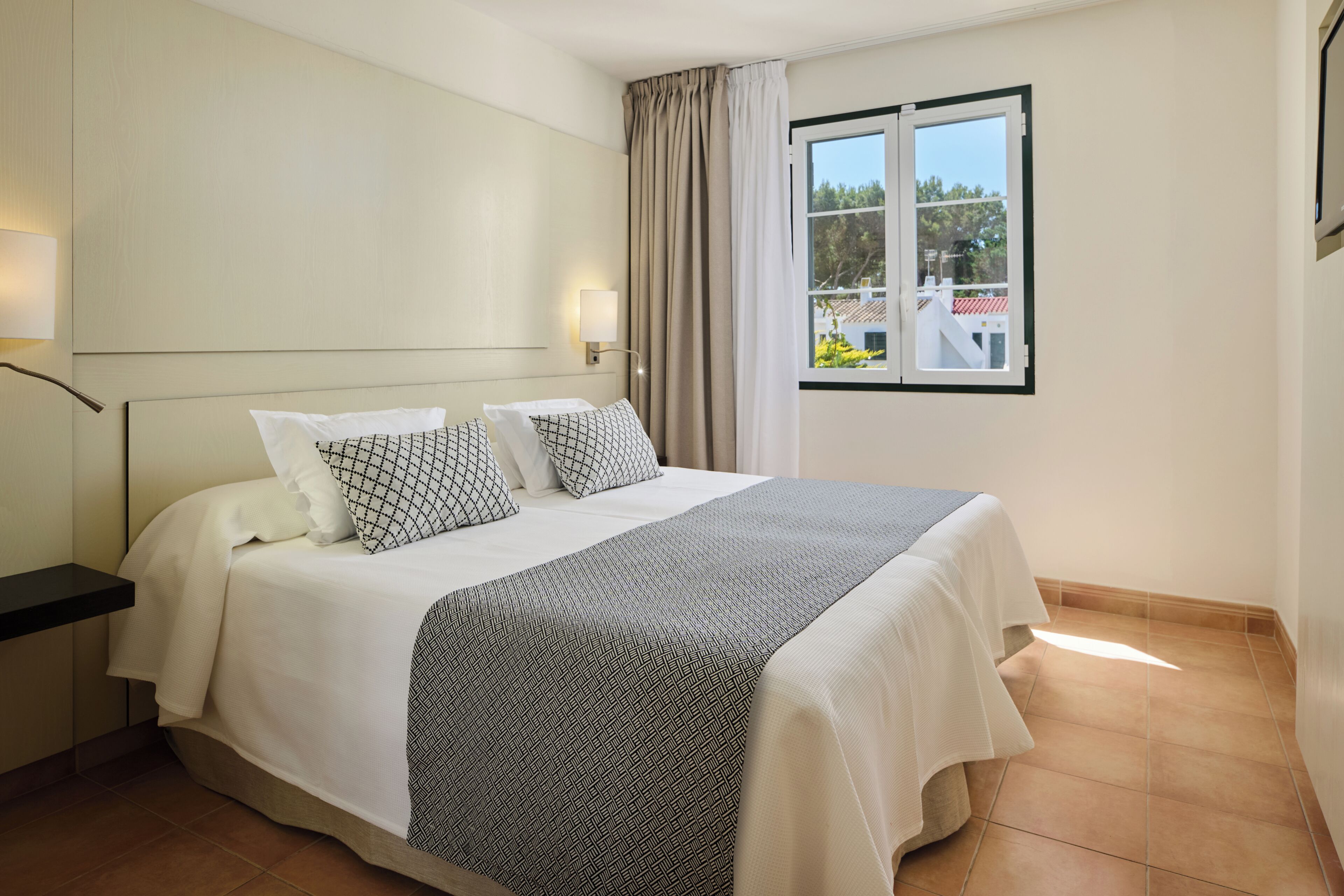 A hotel room with a bed, a nightstand, a lamp, a window with curtains, and a view of the outside.