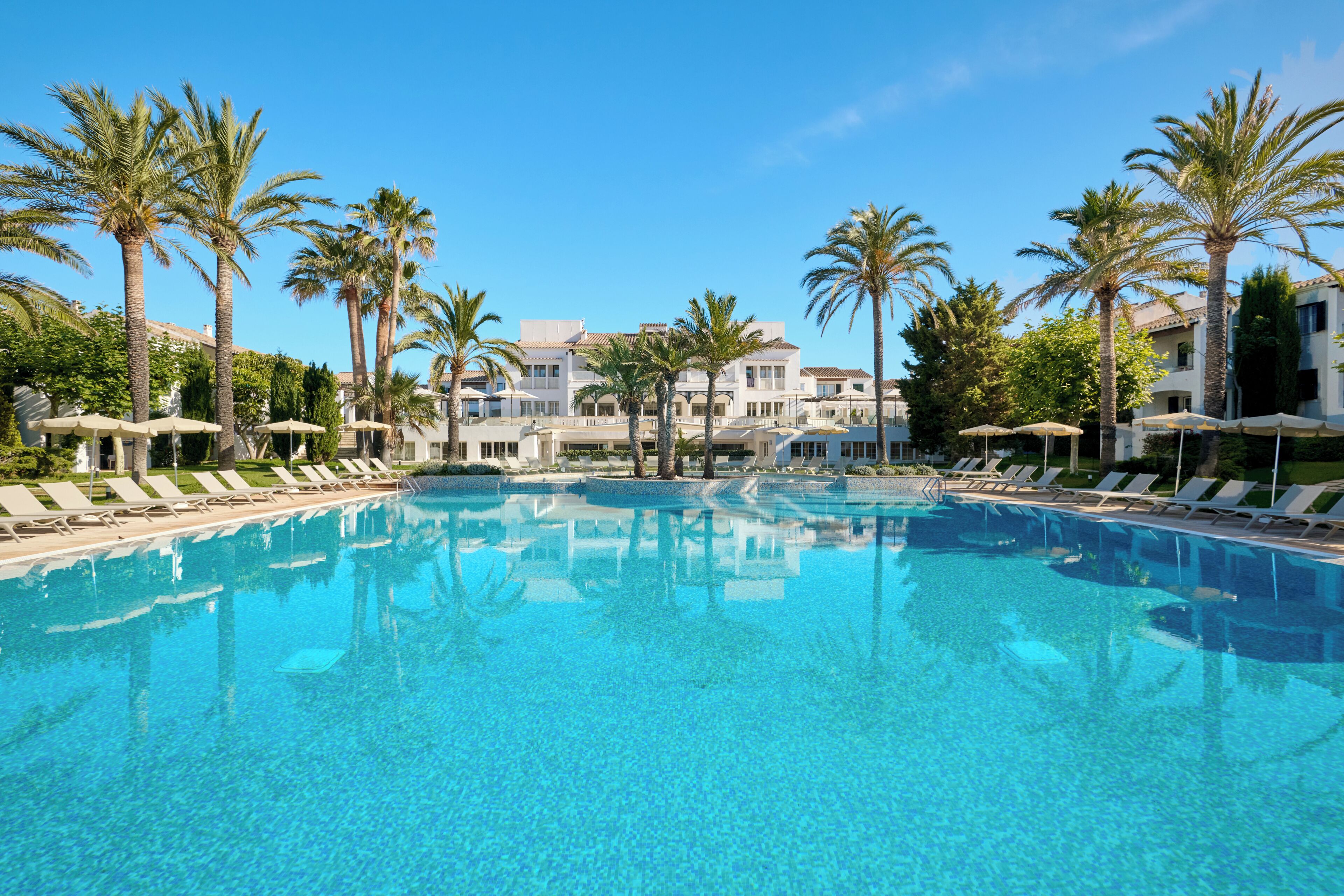 A clear blue pool surrounded by lounge chairs and palm trees, with a building in the background.