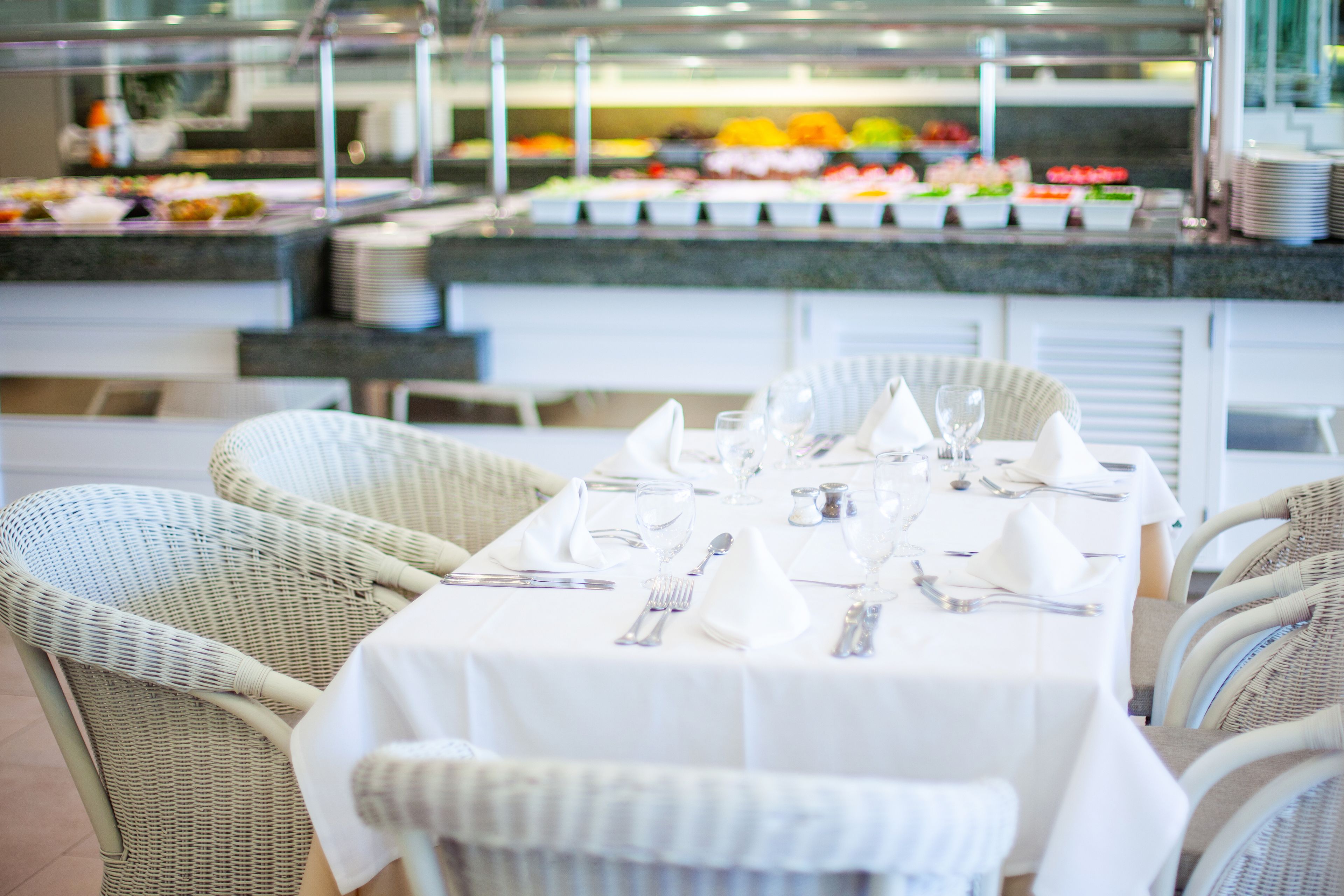 Zona de bufé con diversas opciones de comida, una mesa de comedor preparada para cuatro con mantel blanco y sillas de mimbre, y una barra con platos apilados.