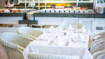 Zona de bufé con diversas opciones de comida, una mesa de comedor preparada para cuatro con mantel blanco y sillas de mimbre, y una barra con platos apilados.