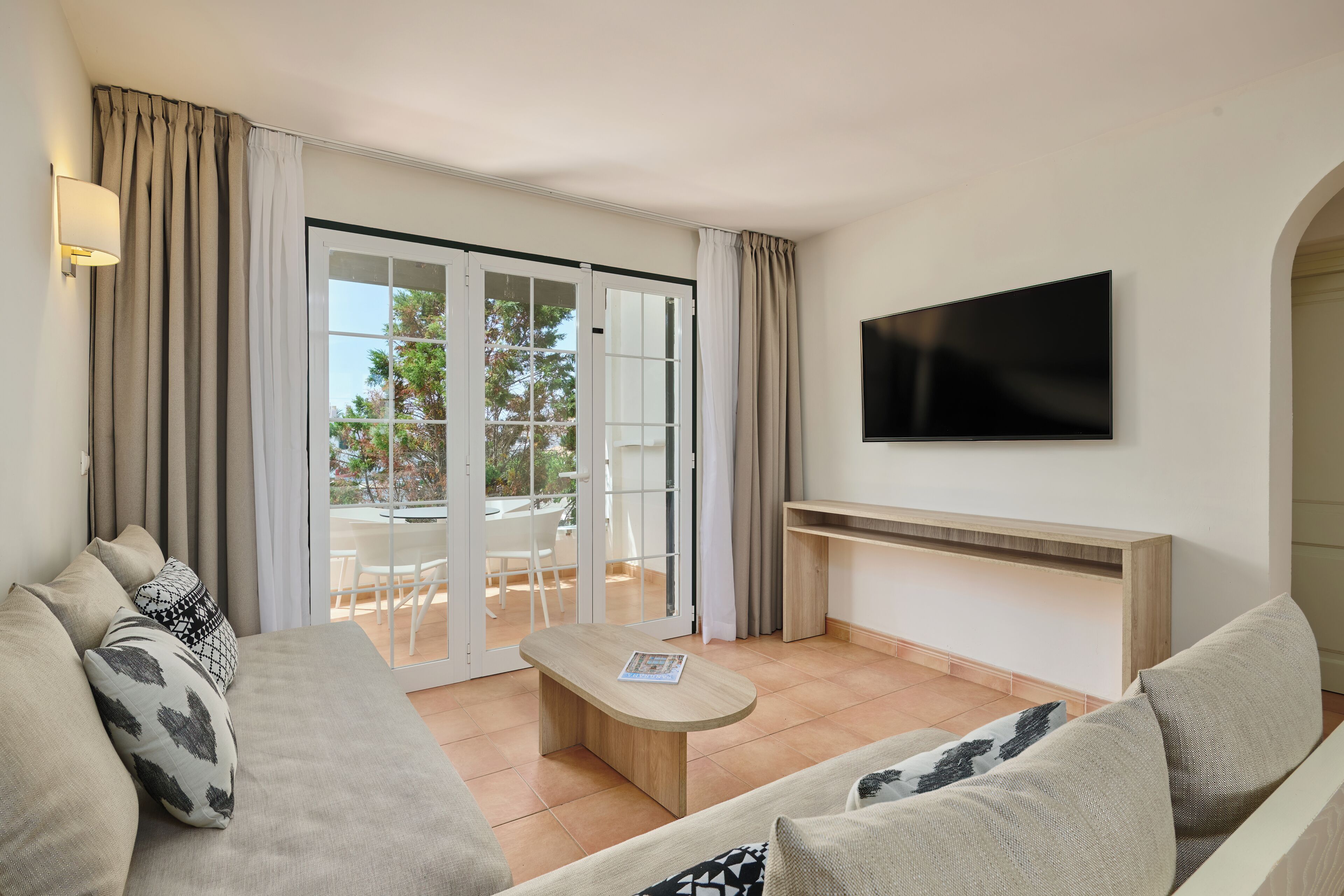 Uma sala de estar moderna com sofá, mesa de centro, televisão de tela plana e uma porta de vidro deslizante que dá para uma área externa com mesa e cadeiras.